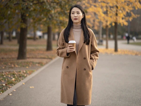 Female nephrologist in a camel coat walking through an autumn park with coffee